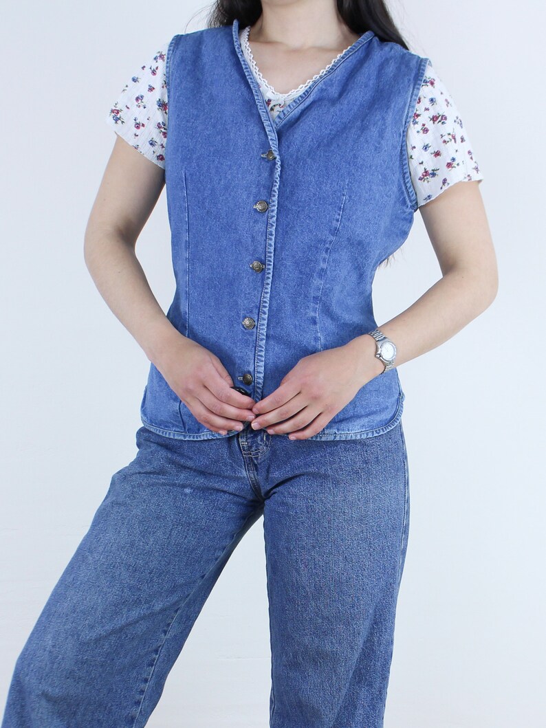 May include: A blue denim vest with a button-up front and a V-neck. The vest is worn over a white floral print shirt and blue jeans.