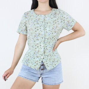 May include: A light green floral print blouse with short sleeves and a button-up front. The blouse features a small, delicate floral pattern in shades of blue, brown, and white. The blouse is paired with denim shorts.