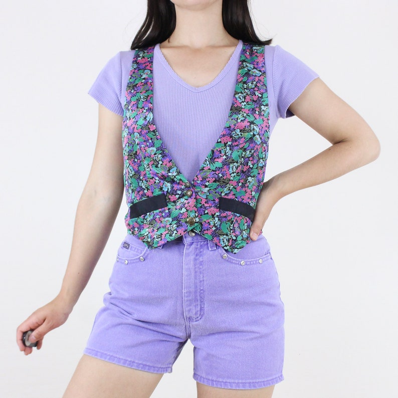May include: A floral print vest with a black trim and a button closure. The vest is worn over a lavender tank top and paired with lavender denim shorts.