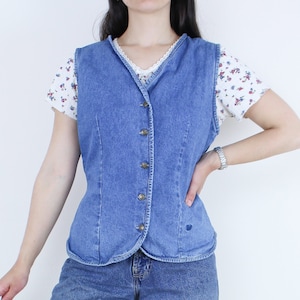May include: A blue denim vest with a button-up front and a pointed collar. The vest is worn over a white shirt with a floral pattern.