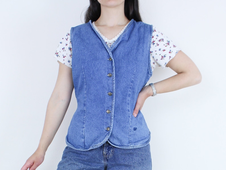 May include: A blue denim vest with a button-up front and a pointed collar. The vest is worn over a white shirt with a floral pattern.