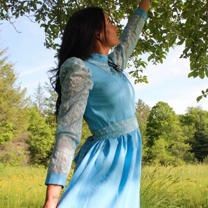 May include: A woman wearing a light blue dress with lace sleeves stands in a field with green grass and trees in the background. She is looking up at the sky.
