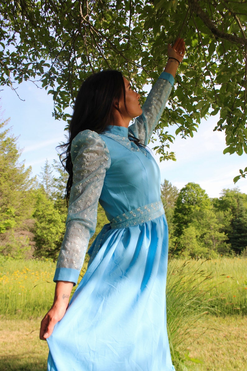 May include: A woman wearing a light blue dress with lace sleeves stands in a field with green grass and trees in the background. She is looking up at the sky.