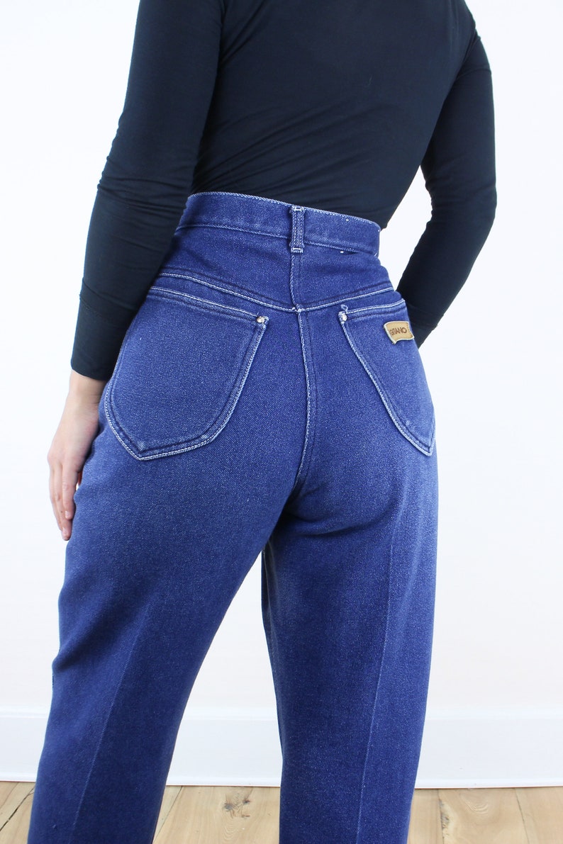 May include: A pair of high-waisted blue denim jeans with a back pocket featuring a brown leather patch with the word "Wrangler" on it.