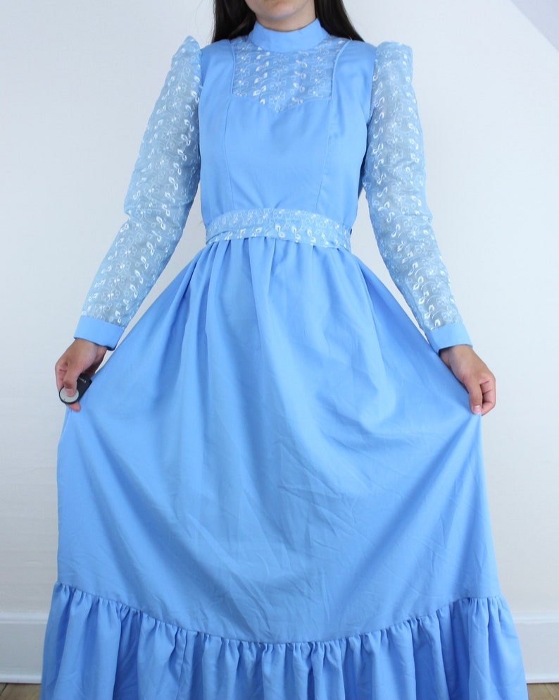 May include: A light blue maxi dress with a high neck and long sleeves. The sleeves are made of a sheer lace fabric with a floral pattern. The dress has a fitted bodice and a full skirt with a ruffle trim.