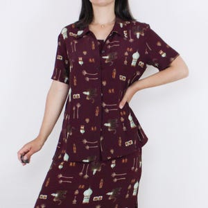 May include: A maroon button-up shirt and matching skirt with a print of stylized animals and geometric shapes.