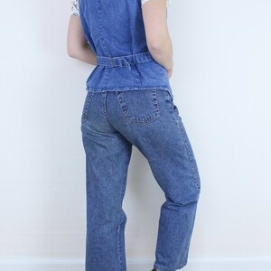 May include: A blue denim vest with a belt and a pair of blue denim jeans. The person is wearing brown clogs.