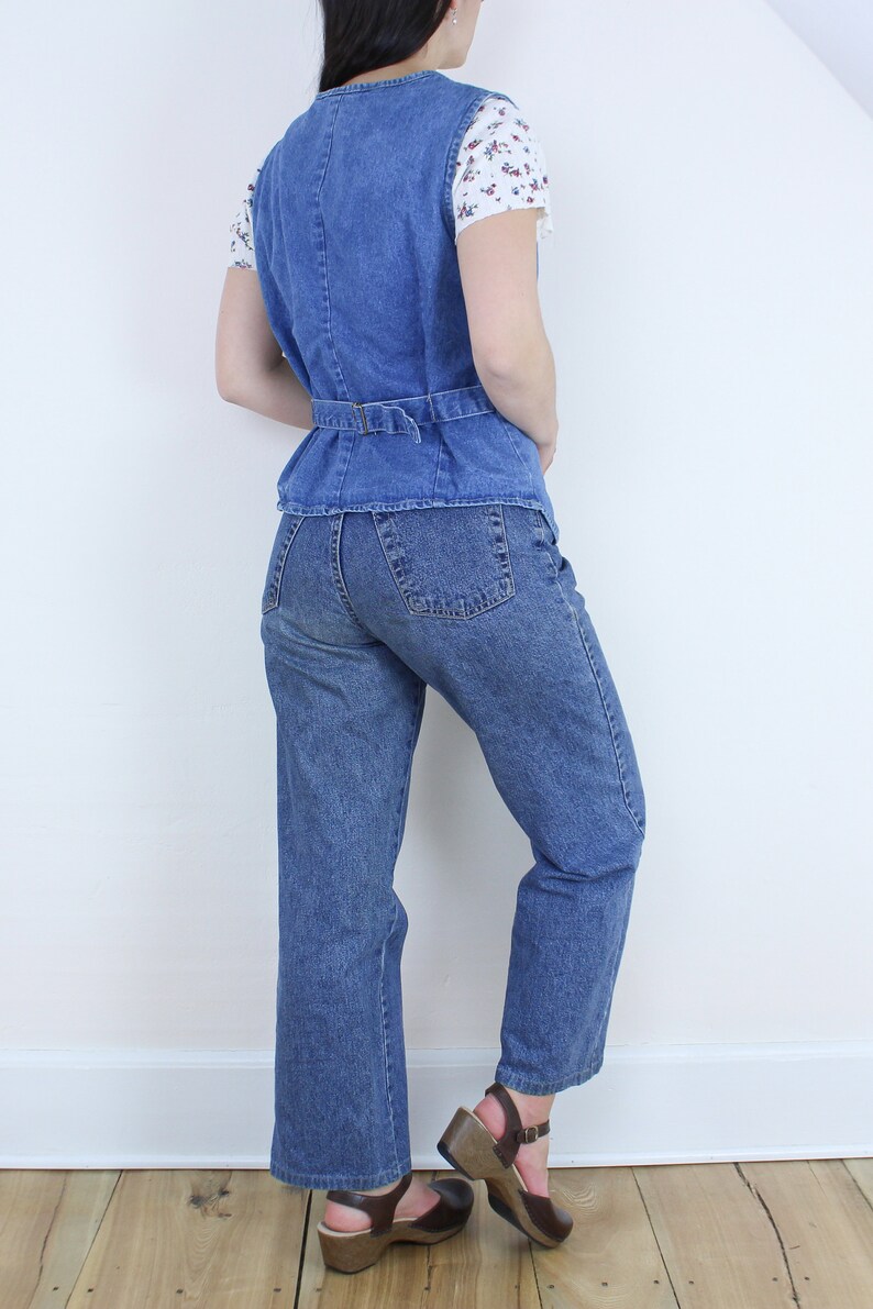 May include: A blue denim vest with a belt and a pair of blue denim jeans. The person is wearing brown clogs.