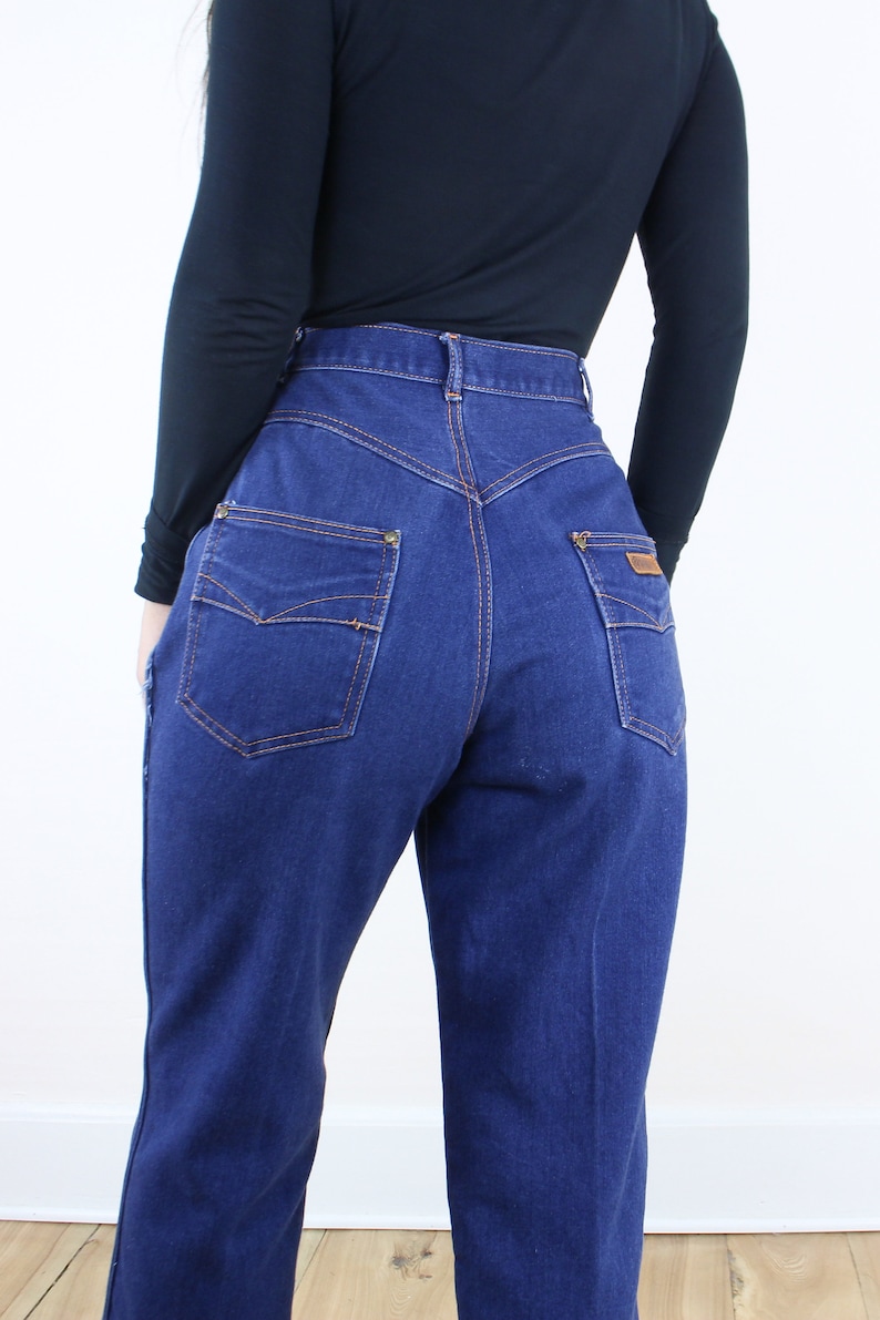 May include: A pair of high-waisted blue denim jeans with a straight leg fit. The jeans have a classic five-pocket design with a brown leather patch on the back right pocket.