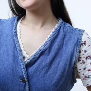 May include: A blue denim vest with a white floral print shirt underneath. The vest has a button closure and a V-neckline.