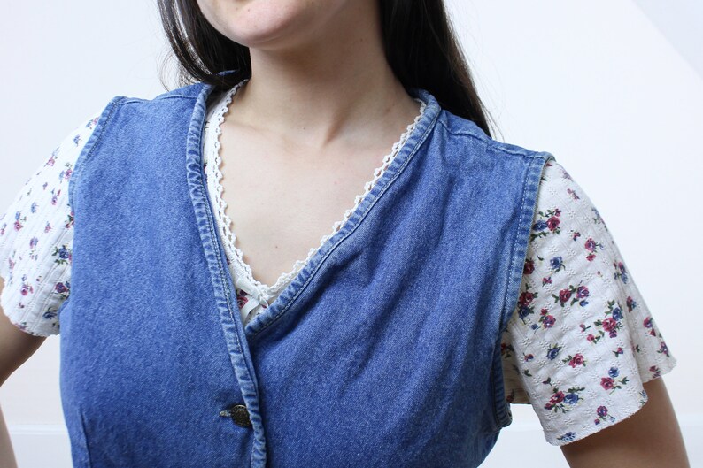 May include: A blue denim vest with a white floral print shirt underneath. The vest has a button closure and a V-neckline.