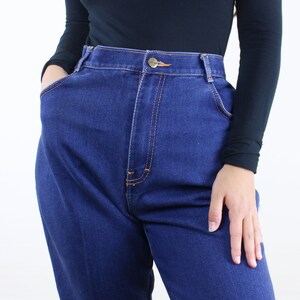 May include: A close-up of a person wearing a black long-sleeved shirt and dark blue high-waisted jeans.