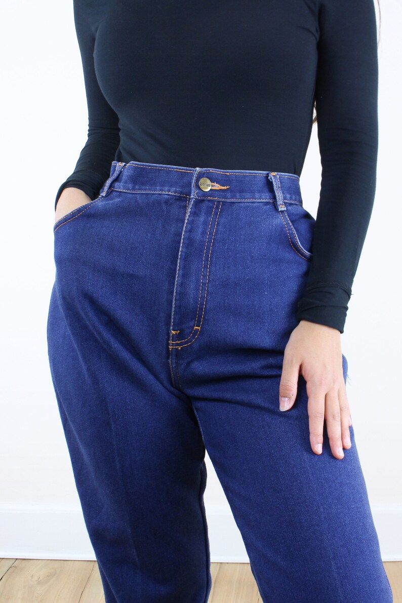 May include: A close-up of a person wearing a black long-sleeved shirt and dark blue high-waisted jeans.