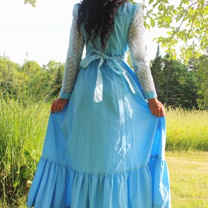 May include: A woman wearing a light blue maxi dress with a white lace overlay and a white ribbon tied at the waist. The dress has a ruffled hem and is worn in a grassy field.