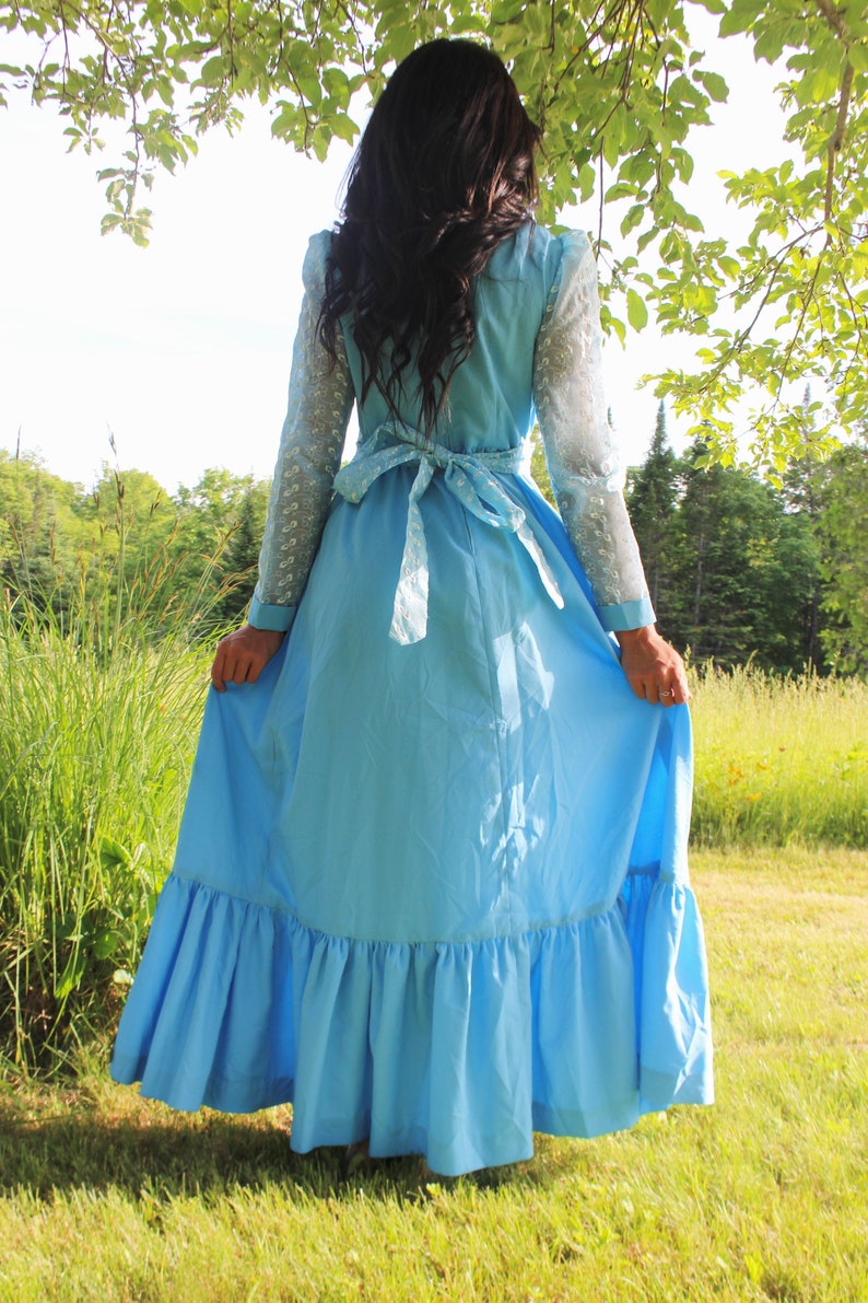 May include: A woman wearing a light blue maxi dress with a white lace overlay and a white ribbon tied at the waist. The dress has a ruffled hem and is worn in a grassy field.