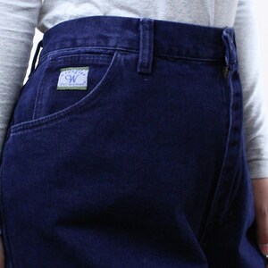 Może przedstawiać: Ciemnoniebieskie dżinsy z denimu z małą zieloną metką z napisem "Wrangler for Woman" na przedniej kieszeni.