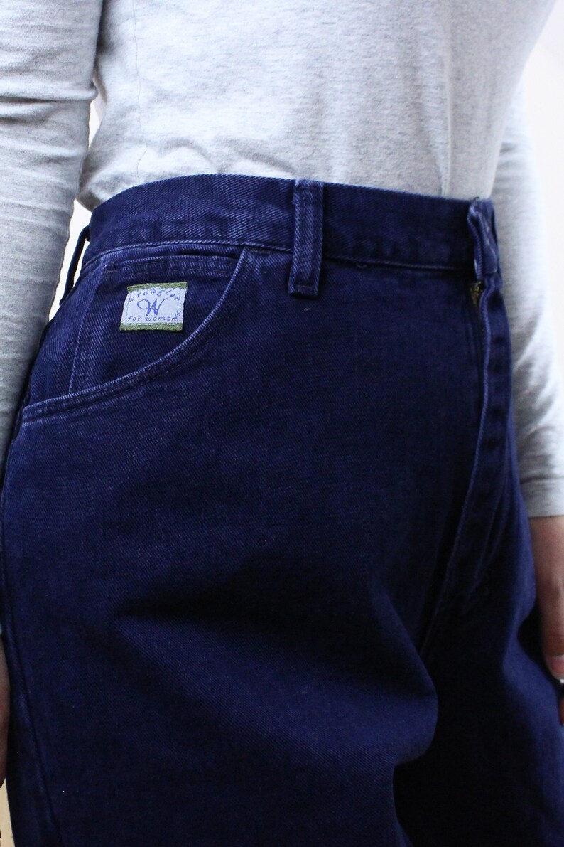 Może przedstawiać: Ciemnoniebieskie dżinsy z denimu z małą zieloną metką z napisem "Wrangler for Woman" na przedniej kieszeni.
