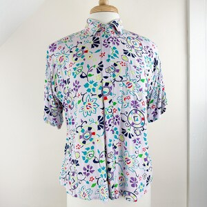 May include: A short-sleeved button-up shirt with a white background and a colorful floral pattern. The pattern includes purple, blue, green, red, and yellow flowers and geometric shapes.