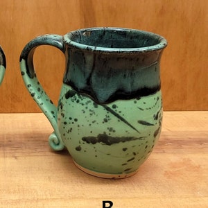 May include: Two ceramic mugs with a green and black glaze. The mugs have a rounded body and a curved handle. The mugs are labeled A and B.