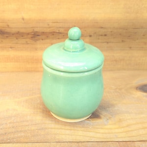 May include: A small, round, light green ceramic jar with a matching lid. The lid features a small, rounded knob handle at the top. The jar has a smooth, glossy finish and a rounded body that tapers slightly toward the base, which shows a light tan color.