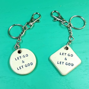 May include: Two keychains with white ceramic charms. One charm is round and says "Let Go & Let God". The other charm is diamond shaped and says "Let Go & Let God".