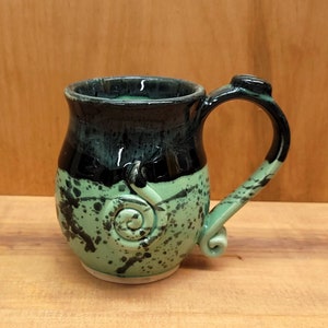 May include: A ceramic mug with a black and green glaze. The mug has a unique swirl design on the front and a handle.