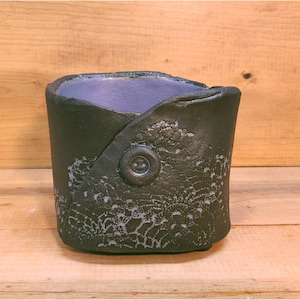 May include: A black ceramic planter with a lace design and a single button on the side. The planter has a purple interior.
