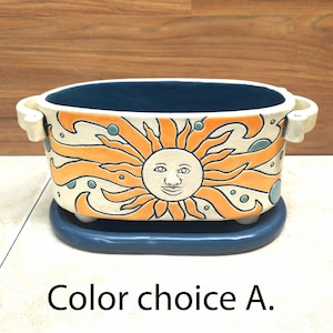 May include: A ceramic planter with a blue rim and a white base. The planter features a sun with a face and orange rays, surrounded by blue and orange swirls. The planter has a matching blue saucer.