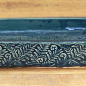 May include: A rectangular ceramic tray with a deep teal glaze and a textured exterior featuring a repeating fern leaf pattern. The interior of the tray is smooth and glossy, while the outer sides display intricate, embossed botanical details.