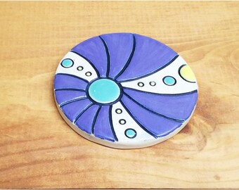 May include: A purple and white ceramic coaster with a swirl design and yellow, green, and blue dots.