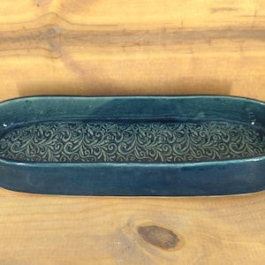 May include: A handmade, oval-shaped ceramic dish in a deep teal color. The interior features an embossed, swirling floral pattern. The dish is displayed on a wooden surface, showcasing its handcrafted nature and decorative design.