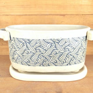 May include: Cream-colored ceramic planter with a blue fern pattern around the middle. The planter has two small handles and sits on a matching saucer. A decorative piece for indoor plants.