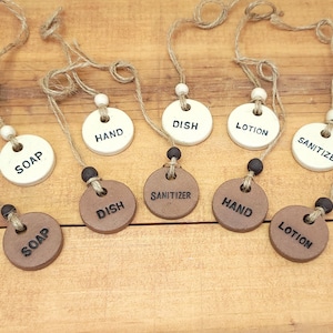 May include: Ceramic soap, dish, lotion, and sanitizer tags with jute twine and wooden beads. The tags are white and brown with black lettering. The tags are hanging on a wooden surface.