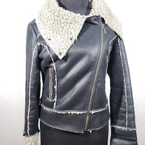 May include: A black faux leather jacket with a cream shearling collar and trim. The jacket has an asymmetrical zip, a small zipped pocket, and shearling cuffs. The jacket is displayed on a mannequin.