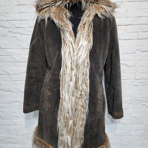 May include: A dark brown suede coat with a hood and faux fur trim. The coat has a long length, with fur trim along the collar, front, and hem. The front has a vertical panel of cream-coloured fur.