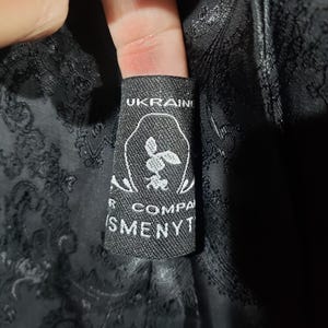 K&ouml;nnte beinhalten: Nahaufnahme eines schwarzen Etiketts mit wei&szlig;em Text und einem floralen Design. Auf dem Etikett steht oben "UKRAINE" und unten "R COMPA" und "SMENYT". Das Etikett ist an einem schwarzen Stoff mit Paisley-Muster befestigt.