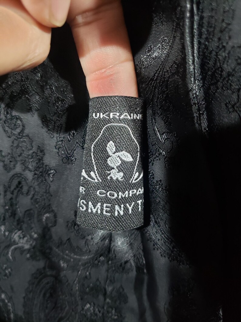 K&ouml;nnte beinhalten: Nahaufnahme eines schwarzen Etiketts mit wei&szlig;em Text und einem floralen Design. Auf dem Etikett steht oben "UKRAINE" und unten "R COMPA" und "SMENYT". Das Etikett ist an einem schwarzen Stoff mit Paisley-Muster befestigt.