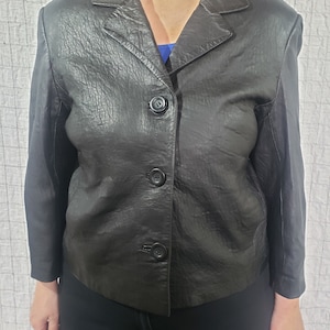 May include: A black leather jacket with a textured finish. The jacket has a notched collar, three black buttons, and long sleeves. The jacket is a classic style.