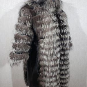 K&ouml;nnte beinhalten: Ein silber-grauer Pelzmantel mit schwarzem Lederinnenfutter. Der Mantel hat ein geschichtetes Design an den &Auml;rmeln und eine lange, flie&szlig;ende Silhouette. Das Fell wirkt weich und luxuri&ouml;s, mit einer Mischung aus hellen und dunklen Graut&ouml;nen.