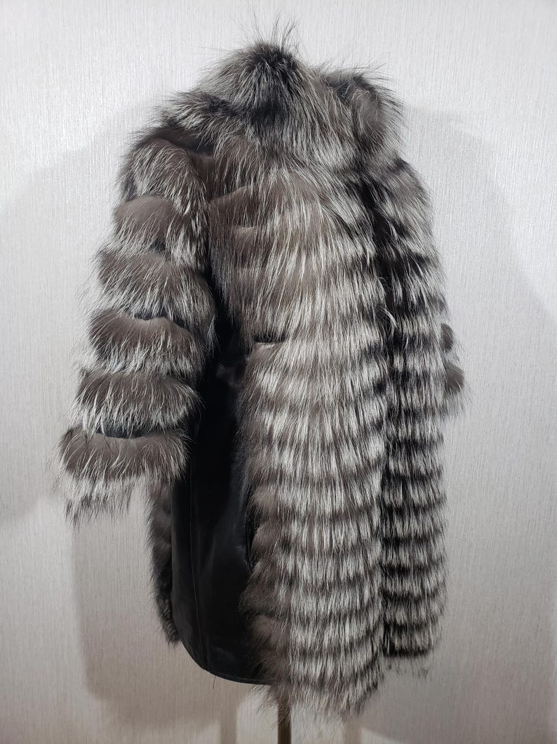 K&ouml;nnte beinhalten: Ein silber-grauer Pelzmantel mit schwarzem Lederinnenfutter. Der Mantel hat ein geschichtetes Design an den &Auml;rmeln und eine lange, flie&szlig;ende Silhouette. Das Fell wirkt weich und luxuri&ouml;s, mit einer Mischung aus hellen und dunklen Graut&ouml;nen.