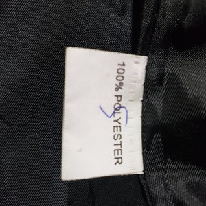 以下が含まれることがあります： 黒い生地を背景に、黒い文字で「100% POLYESTER」と書かれた白いタグ。青いマークが付いています。