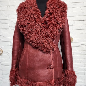 Könnte beinhalten: Burgunderfarbene Lederjacke mit großem, lockigem, rötlich-braunem Shearling-Kragen, Manschetten und Saum. Die Jacke hat einen Knopfverschluss und wird auf einer schwarzen Schaufensterpuppe präsentiert. Die Jacke ist ein stilvolles Oberbekleidungsstück.