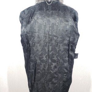 K&ouml;nnte beinhalten: Schwarze Jacke mit grauem Pelzkragen und -besatz. Das Innenfutter hat ein Paisley-Muster. Eine silberne Kette ist am Kragen befestigt. Die Jacke wird von hinten gezeigt.