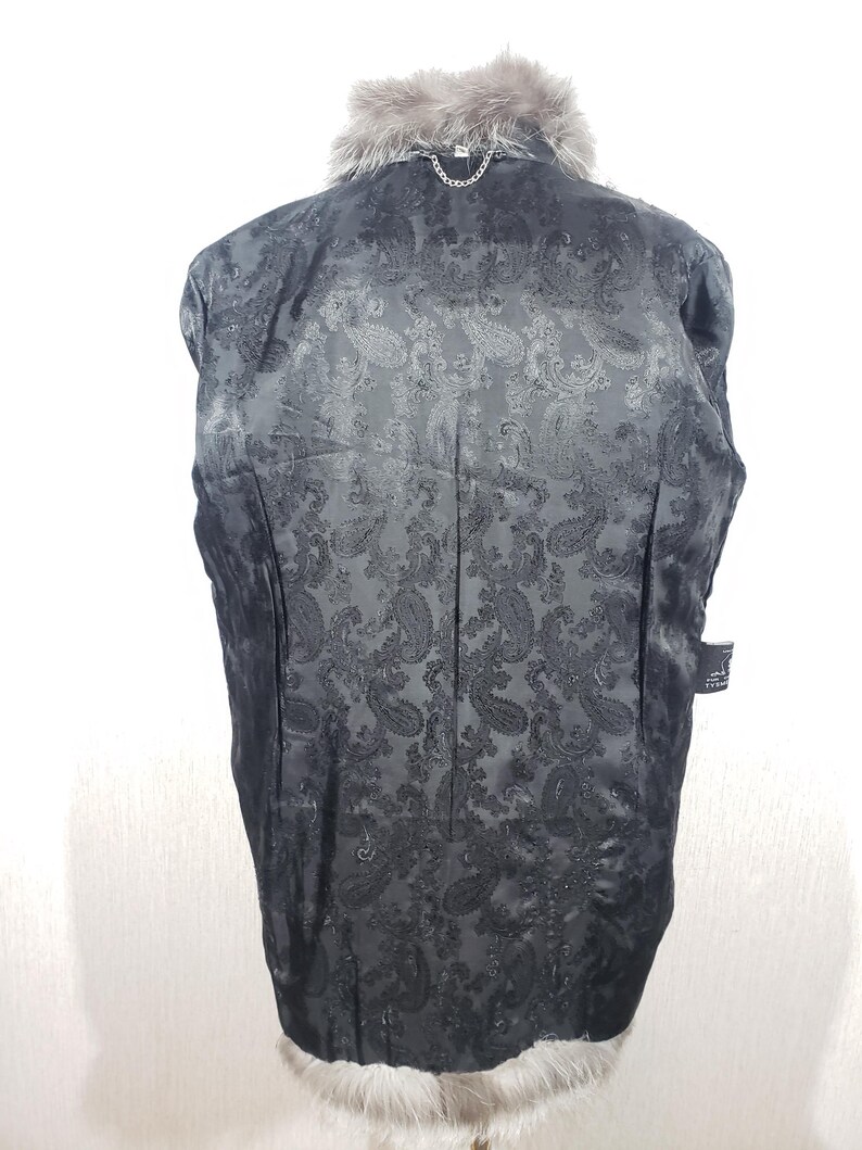 K&ouml;nnte beinhalten: Schwarze Jacke mit grauem Pelzkragen und -besatz. Das Innenfutter hat ein Paisley-Muster. Eine silberne Kette ist am Kragen befestigt. Die Jacke wird von hinten gezeigt.
