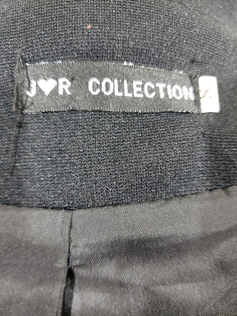 以下が含まれることがあります： 「J♡R COLLECTION」と書かれたラベルが付いた黒い衣服のクローズアップ。 ラベルは長方形で、白い文字とハートのシンボルが付いています。 衣服は、テクスチャード加工された生地で作られているようです。