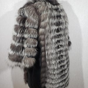 K&ouml;nnte beinhalten: Ein luxuri&ouml;ser Silberfuchsfellmantel. Der Mantel hat eine Mischung aus silbernem, grauem und schwarzem Fell mit einem weichen, strukturierten Aussehen. Das Design umfasst einen gro&szlig;en Kragen und &Auml;rmel mit einem geschichteten, strukturierten Muster. Der Mantel ist ein Statement-Piece.