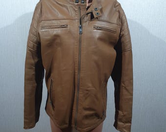 Può includere: Giacca in pelle marrone con zip, due tasche sul petto e due tasche laterali. La giacca ha un colletto rialzato e dettagli trapuntati sulle spalle. La giacca è esposta su un manichino.