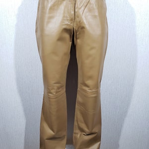 May include: Tan leather trousers with a button fastening, pockets, and a subtle flare at the hem. The trousers are shown on a mannequin against a plain white backdrop. The leather has a smooth, matte appearance.