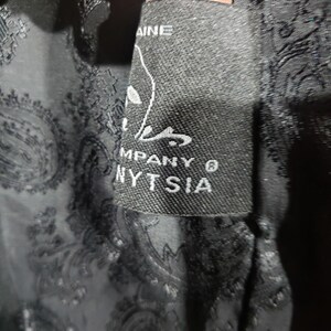 K&ouml;nnte beinhalten: Nahaufnahme eines schwarzen Etiketts mit wei&szlig;em Text und einer stilisierten Grafik. Das Etikett lautet "NYTSIA COMPANY &reg;" und ist in einen schwarzen Paisley-Stoff eingen&auml;ht. Der Stoff hat ein subtiles, sich wiederholendes Muster.