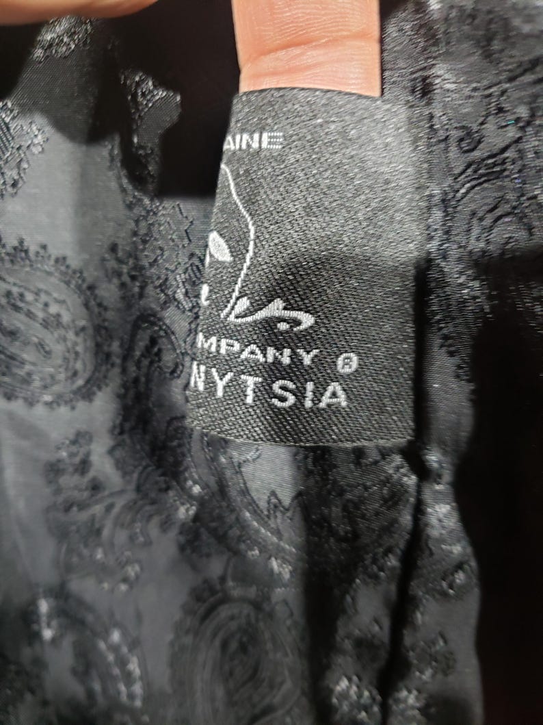 K&ouml;nnte beinhalten: Nahaufnahme eines schwarzen Etiketts mit wei&szlig;em Text und einer stilisierten Grafik. Das Etikett lautet "NYTSIA COMPANY &reg;" und ist in einen schwarzen Paisley-Stoff eingen&auml;ht. Der Stoff hat ein subtiles, sich wiederholendes Muster.